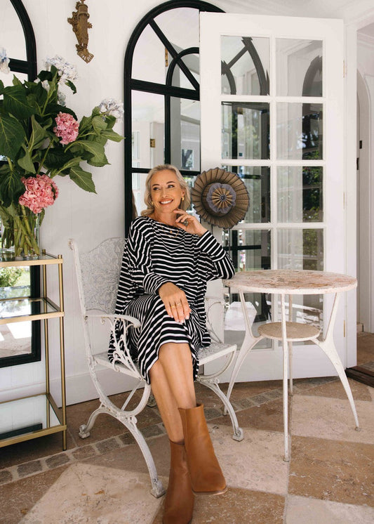 A woman wearing the Susie Oversized Asymmetrical Bamboo Top in black & white stripes and brown boots sits on a white chair at a small round table in a bright room with large windows and flowers, smiling and looking to the side.