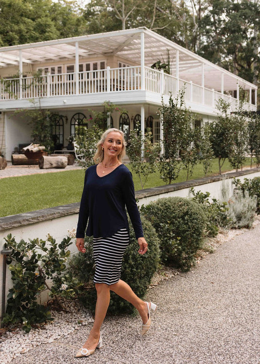 A smiling woman with long blonde hair, wearing the Sia V Neck Bamboo Longsleeve Top in Navy and a striped skirt, walks along a gravel path in front of a large white house with a wraparound porch and lush green landscaping.