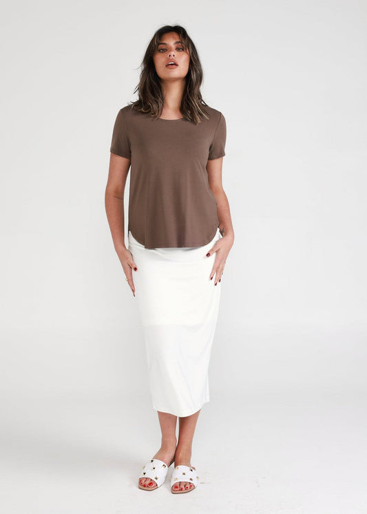 A woman with shoulder-length hair and a neutral expression stands against a white background, wearing the Janis Short Sleeve Bamboo T-Shirt in Chocolate, paired with a white midi skirt and white sandals.