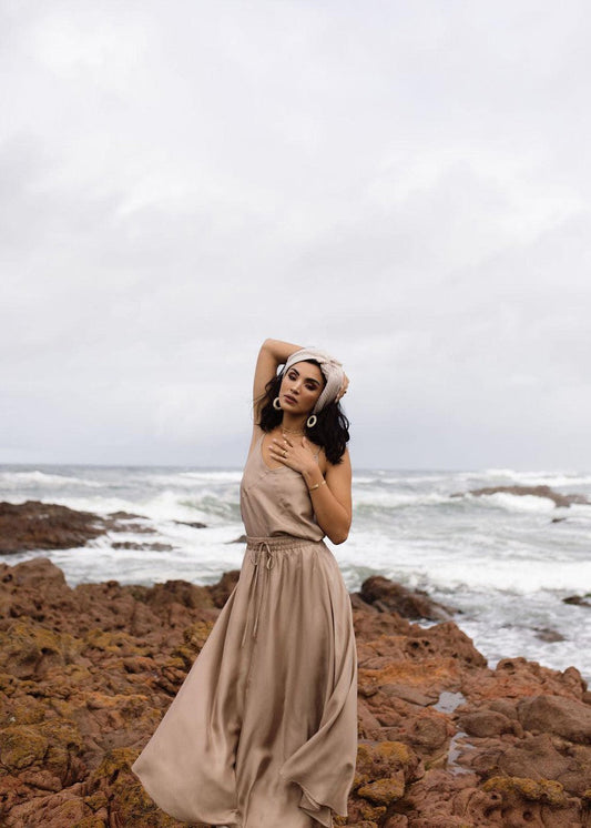 A woman stands on a rocky shore under a cloudy sky, wearing the Aster Silk Maxi Skirt in Bronze and a white headscarf, posing with one hand on her head and the other on her chest as waves crash behind her.
