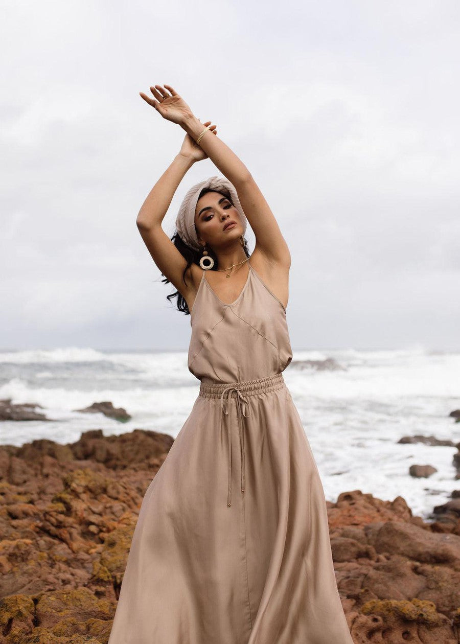 A woman in a light beige dress and headscarf stands on rocky terrain by the ocean, arms raised as waves crash behind her, embodying effortless style in the Clover Sleeveless Silk Cami - Bronze against a cloudy sky.