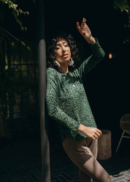 Wearing large earrings and beige pants, a woman with curly hair stands outdoors in sunlight, one arm raised and eyes closed, dressed in the Adele Long Sleeve Bamboo Top - Green Leopard.