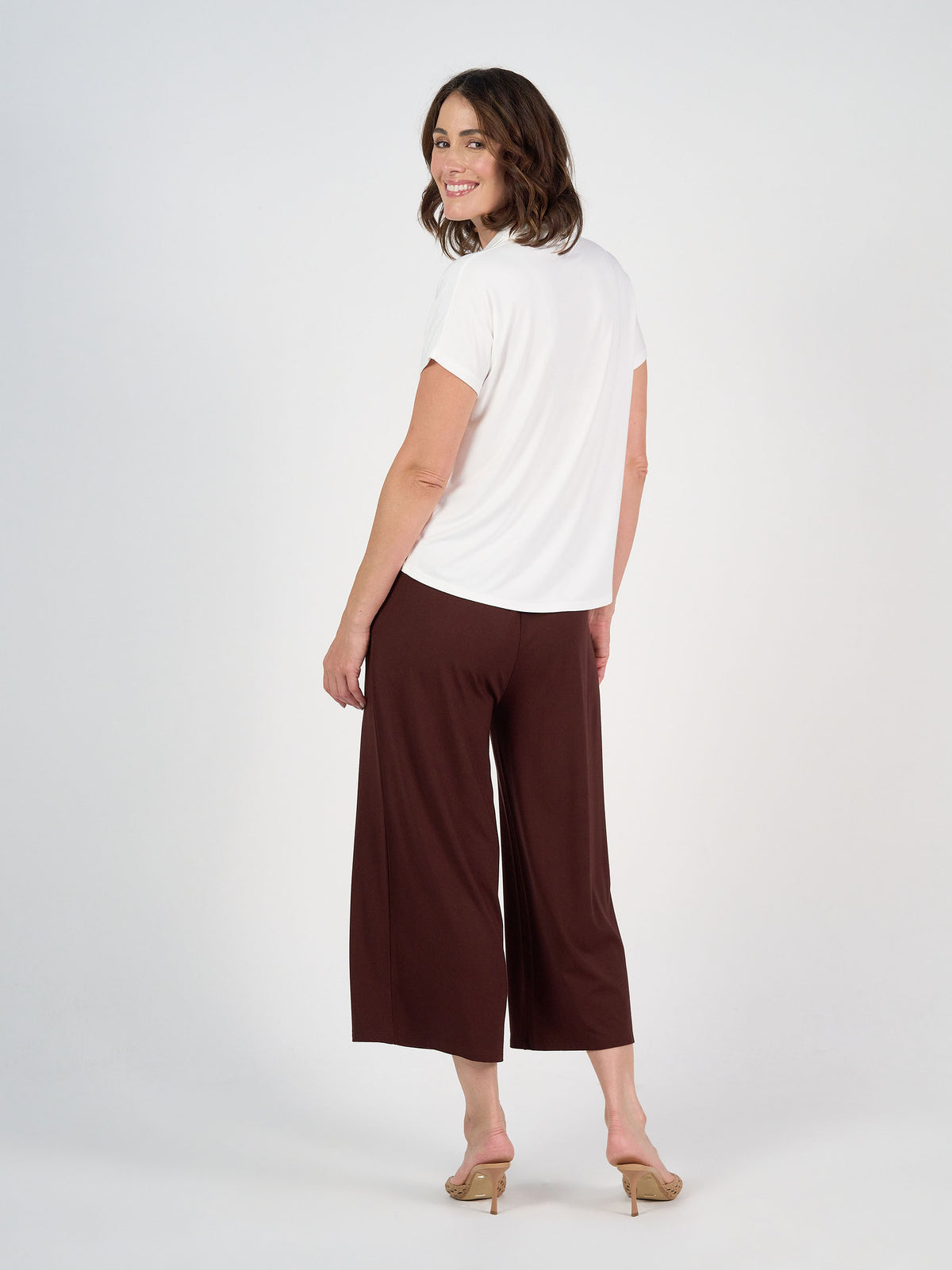 A woman stands against a plain light background, looking back over her shoulder, wearing the Rush Bamboo Short Sleeve Collared Polo Top in cream, paired with brown wide-leg pants and beige heels.