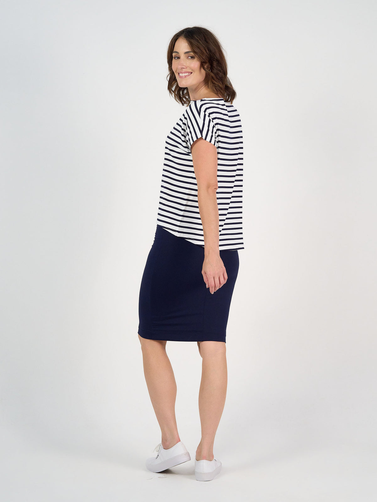 A woman stands sideways, looking back and smiling. She wears the Wilde Bamboo Box Fit Tee in navy & white stripe, paired with a navy knee-length skirt and white sneakers against a plain white background.