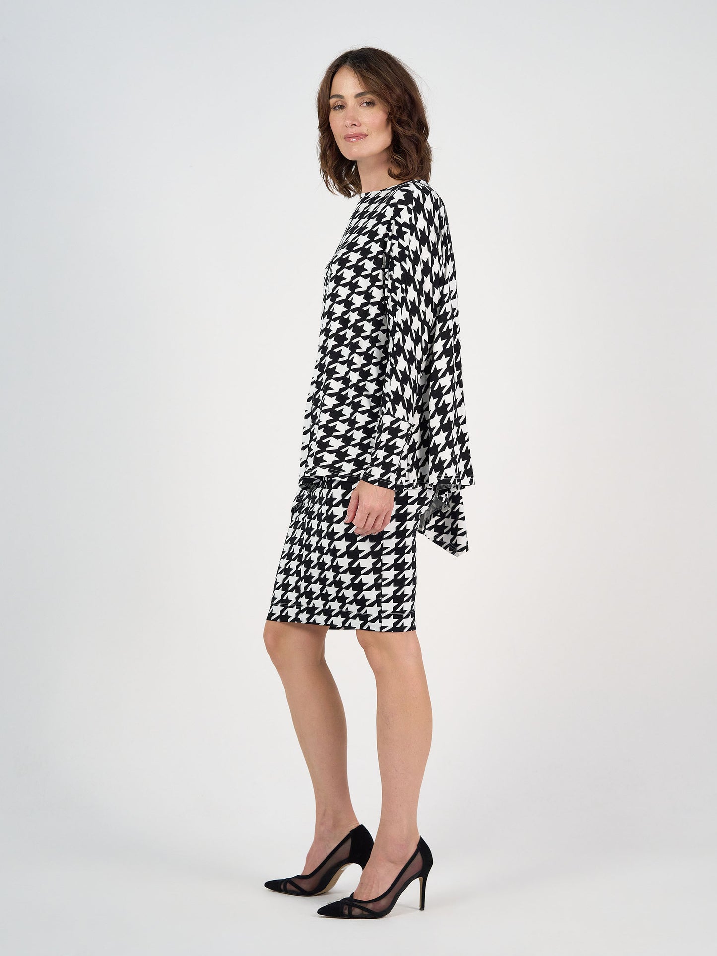 A woman stands against a plain background wearing the Susie Oversized Asymmetrical Bamboo Top in houndstooth, styled as a knee-length dress with long sleeves and black heels. She faces sideways, glancing toward the camera.