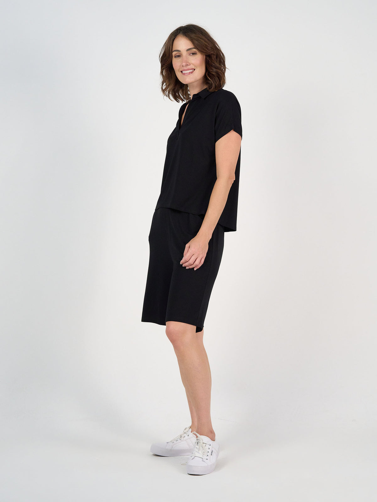 A woman with shoulder-length brown hair smiles sideways, wearing the Rush Bamboo Short Sleeve Collared Polo in black with matching knee-length shorts and white sneakers, against a plain white background.