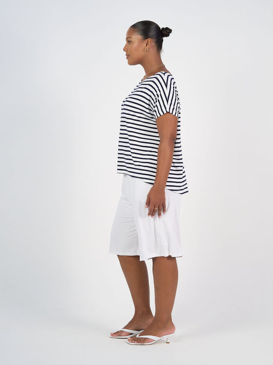 A woman stands in profile against a white background, wearing the Wilde Bamboo Box Fit Tee in navy & white stripe, white knee-length shorts, white heeled sandals, and gold hoop earrings. Her hair is tied back in a neat bun.