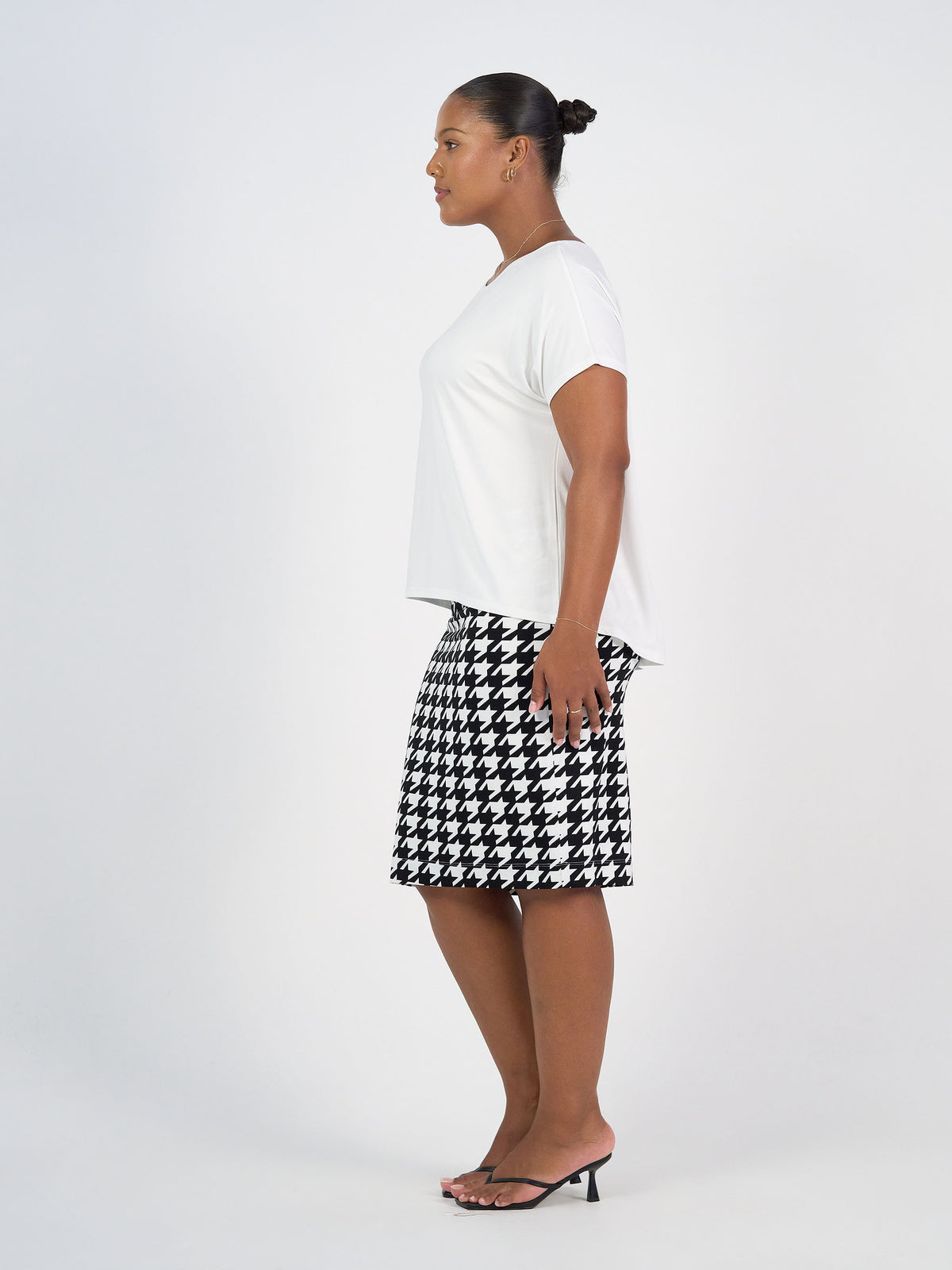 A woman in profile against a white background wears the Wilde Bamboo Box Fit Tee in cream, paired with a black and white houndstooth skirt and black heeled sandals. Her hair is styled in a neat bun.