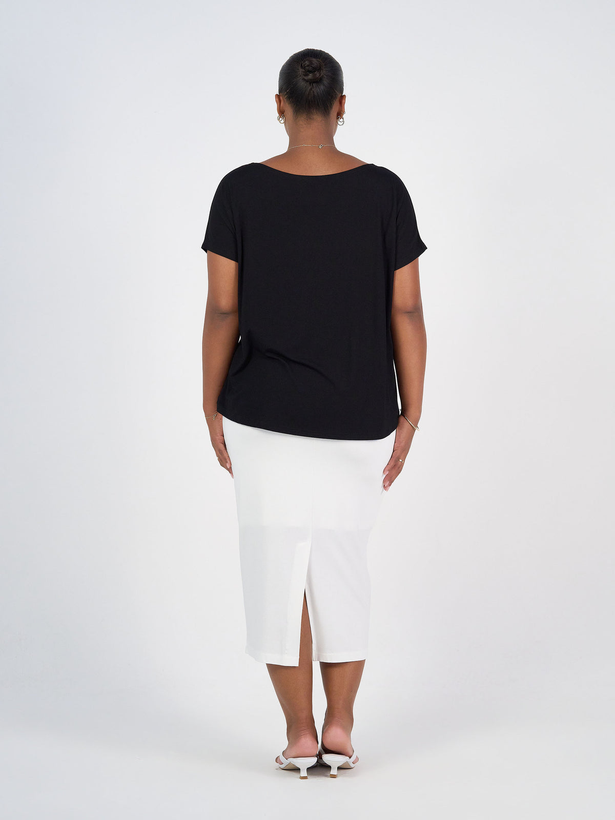 A woman stands with her back to the camera, wearing the Wilde Bamboo Box Fit Tee in black, a white knee-length skirt with a back slit, and open-toe heels against a plain white background.