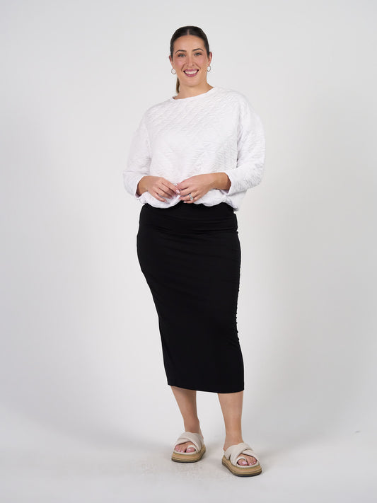 A woman smiles against a white background, wearing the Sacha Oversized Quilted Bamboo Sweater in white, a fitted black midi skirt, beige slide sandals, hoop earrings, and her hair pulled back.