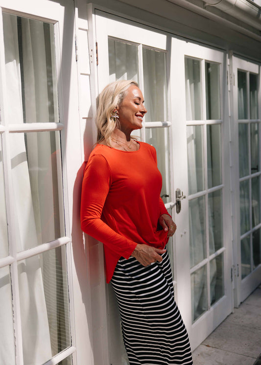 A woman with blonde hair smiles while standing by glass doors, wearing the Adele Long Sleeve Bamboo Top in Fiery Red and a black-and-white striped skirt, as sunlight casts gentle shadows on the scene.