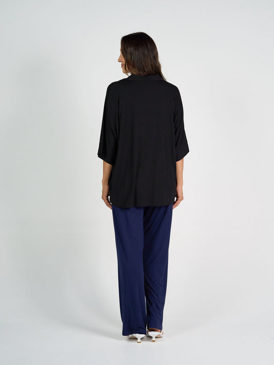 A woman faces away, wearing the Alanis 3/4 Sleeve Collared Bamboo Shirt in black with wide-leg navy pants and white heeled sandals, set against a plain light background.