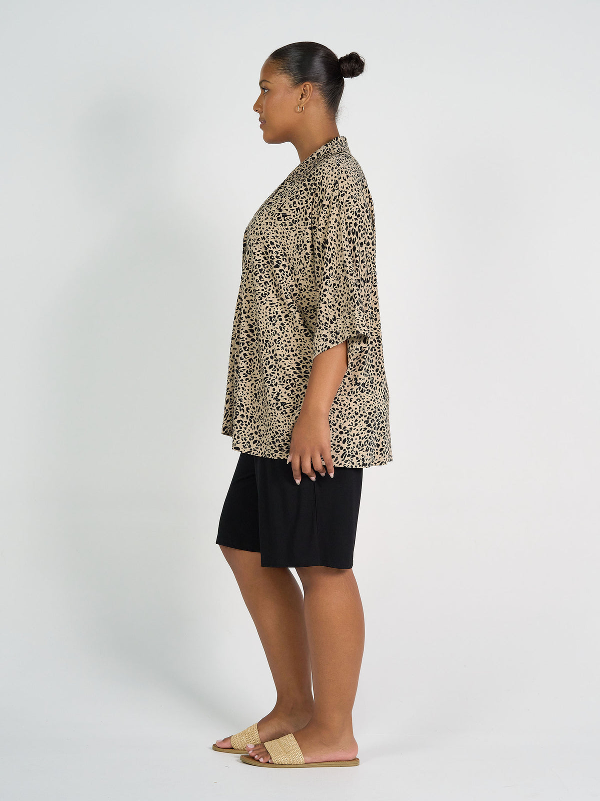 A woman stands in profile against a white background, wearing the Alanis 3/4 Sleeve Collared Shirt in brown leopard, paired with black knee-length shorts and beige slide sandals. Her hair is pulled back in a neat bun.