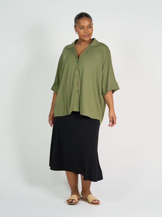 A woman stands against a white background wearing the Alanis 3/4 Sleeve Collared Bamboo Shirt in olive, paired with a black midi skirt and beige flat sandals. Her hair is pulled back, and she poses with a relaxed, confident stance.