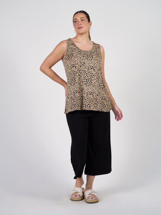 A woman poses against a plain background wearing the Alicia Reversible Bamboo Singlet Top in Brown Leopard, loose black cropped pants, and white platform sandals. Her hair is tied back and she stands with one hand on her hip.