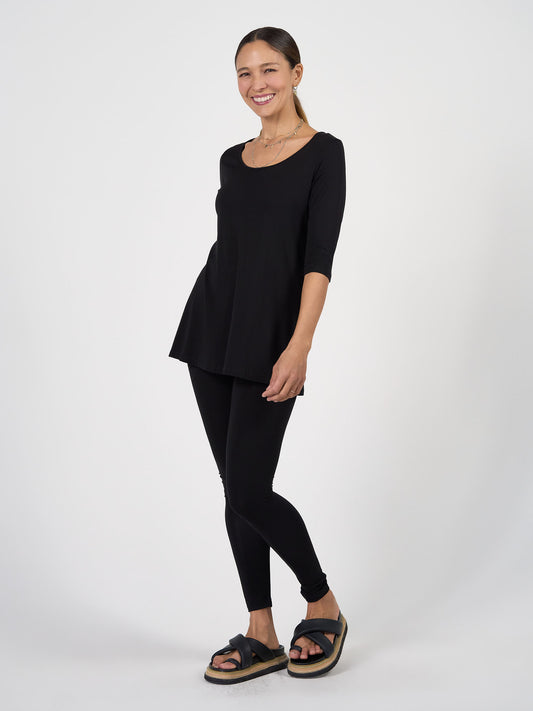 A woman with tied-back hair smiles against a white background, striking a relaxed pose in a black tunic top, Luxe Bamboo Full-Length Legging - Black, black platform sandals, and a gold necklace.