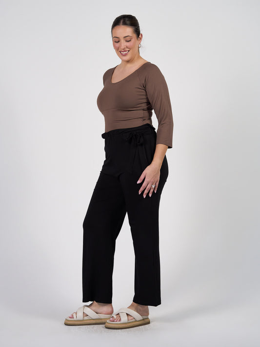 A woman stands smiling against a white background, wearing the Ava 3/4 Sleeve Cropped Bamboo Top in brown as a versatile layer, paired with high-waisted black pants with a tied belt and white slide sandals.