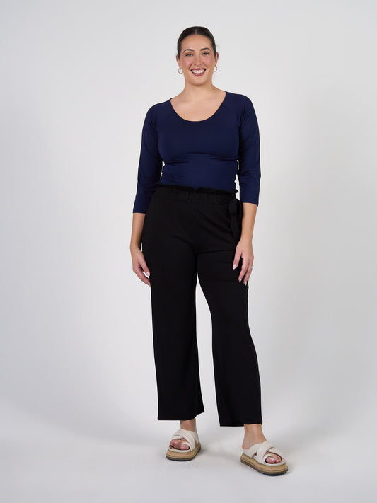A woman stands smiling against a white background, wearing the Ava 3/4 Sleeve Cropped Bamboo Top in navy blue, paired with black wide-leg pants, beige sandals, her hair tied back, and hoop earrings.