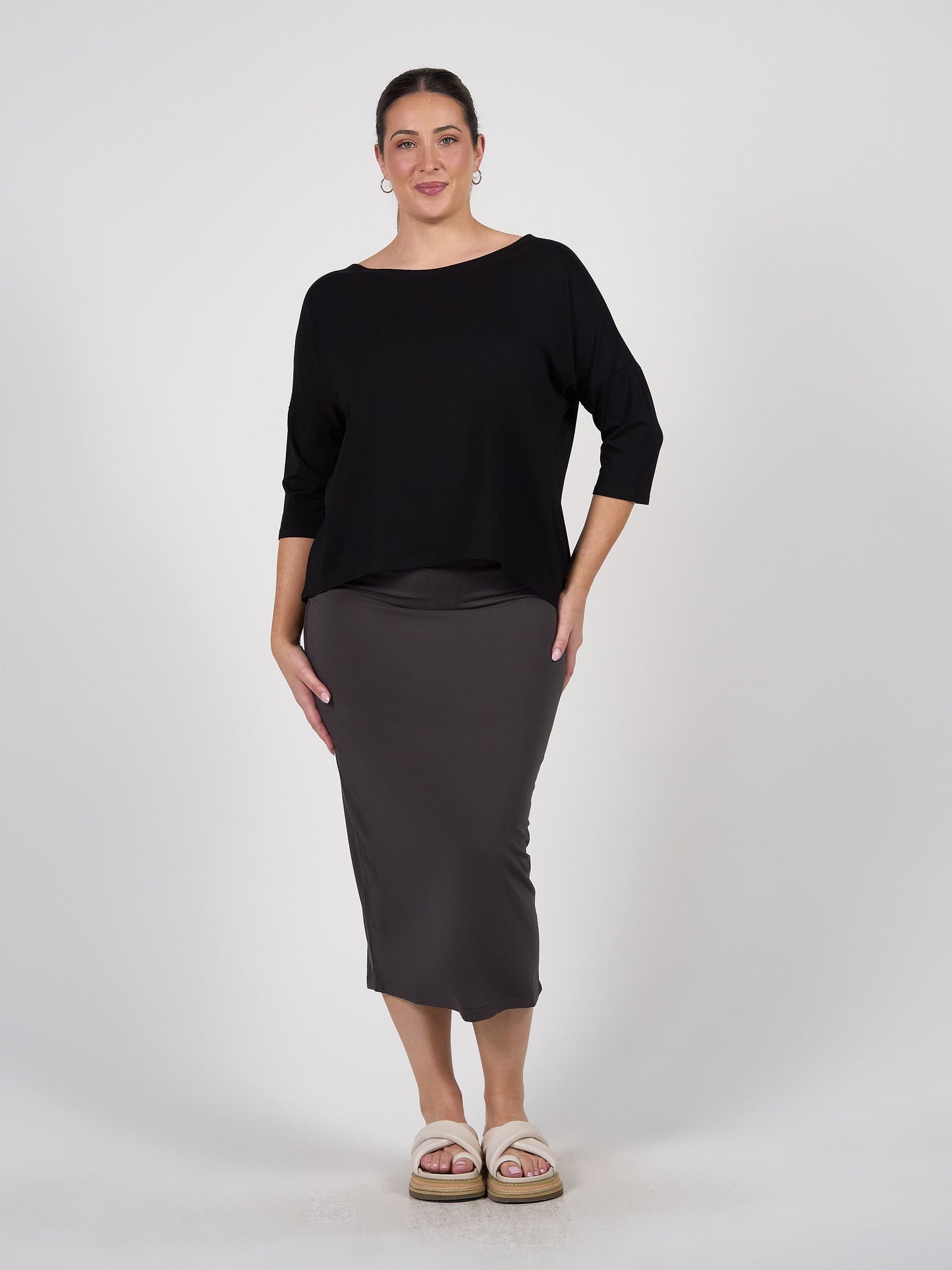 A woman, smiling with her hair pulled back, stands against a white background wearing the Camila 3/4 Sleeve Bamboo Top in black, paired with a fitted gray midi skirt and light slip-on sandals.