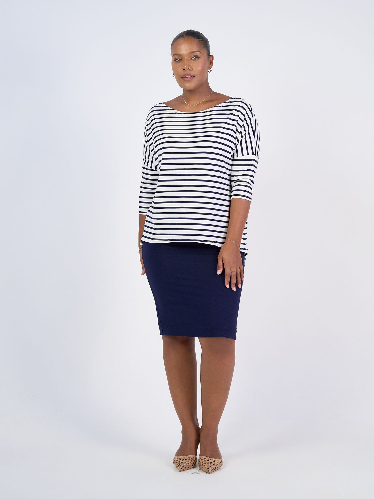 A woman stands against a white background, wearing the Camila 3/4 Sleeve Bamboo Top in Navy & White Stripe, paired with a navy knee-length skirt and beige pointed-toe shoes. She looks forward with a neutral expression.