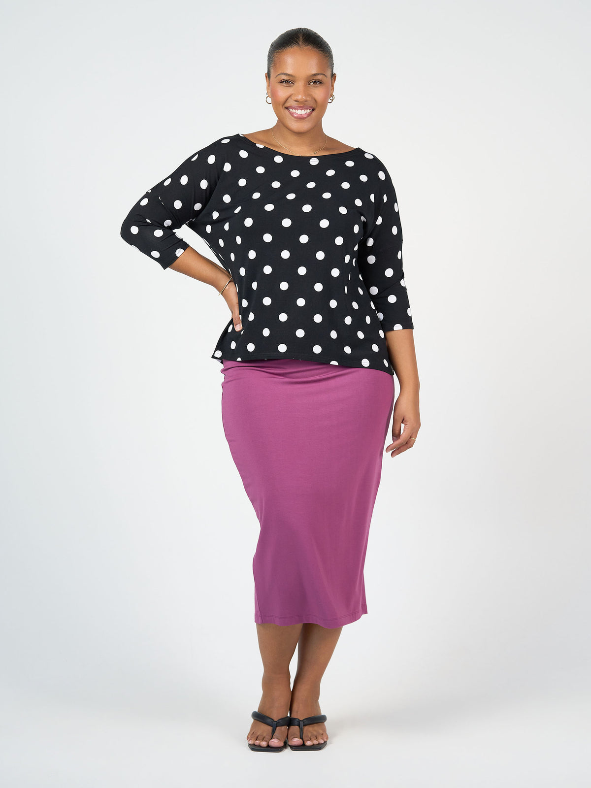 A smiling woman with one hand on her hip wears the Camila 3/4 Sleeve Bamboo Top - Polka Dot, paired with a purple midi skirt and black sandals, standing against a plain light background.