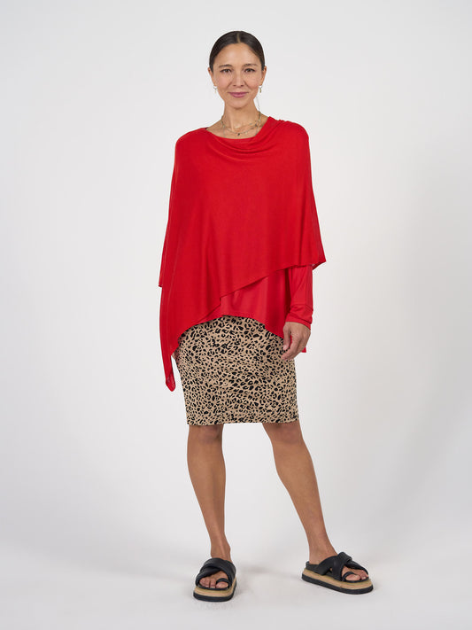A woman with tied-back hair smiles gently, modeling the Carrie Cashmere & Bamboo Knit Poncho in bright red over a leopard print skirt and black platform sandals, standing against a plain white background.
