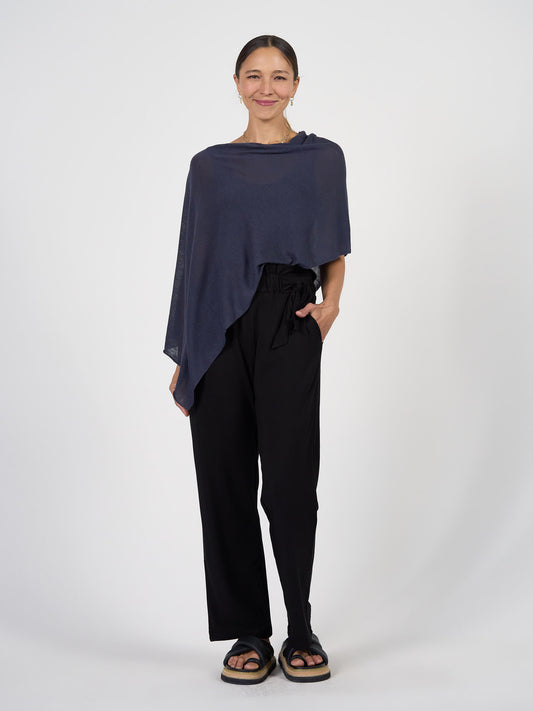 A woman stands against a white background, wearing the Carrie Cashmere & Bamboo Knit Poncho in dark blue, paired with black high-waisted pants and black platform sandals. Her hair is pulled back, one hand in her pocket, and she's smiling.