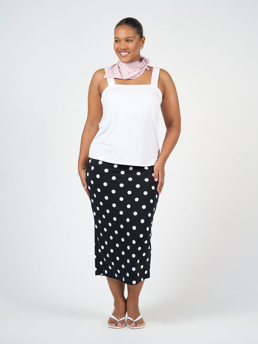 A woman smiles while wearing a white tank top, pale pink scarf, white sandals, and the Maxi Bamboo Whitney Tube Skirt - Polka Dot, standing against a plain light background.