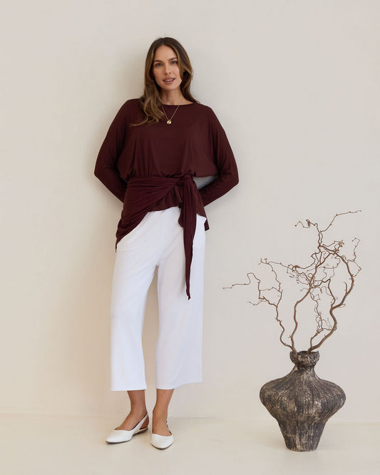 A woman stands against a neutral wall wearing a long-sleeve maroon wrap top, white wide-leg pants, and flats. She completes her look with the Sassoon Cashmere/Bamboo Scarf, standing beside a large decorative vase with bare branches.