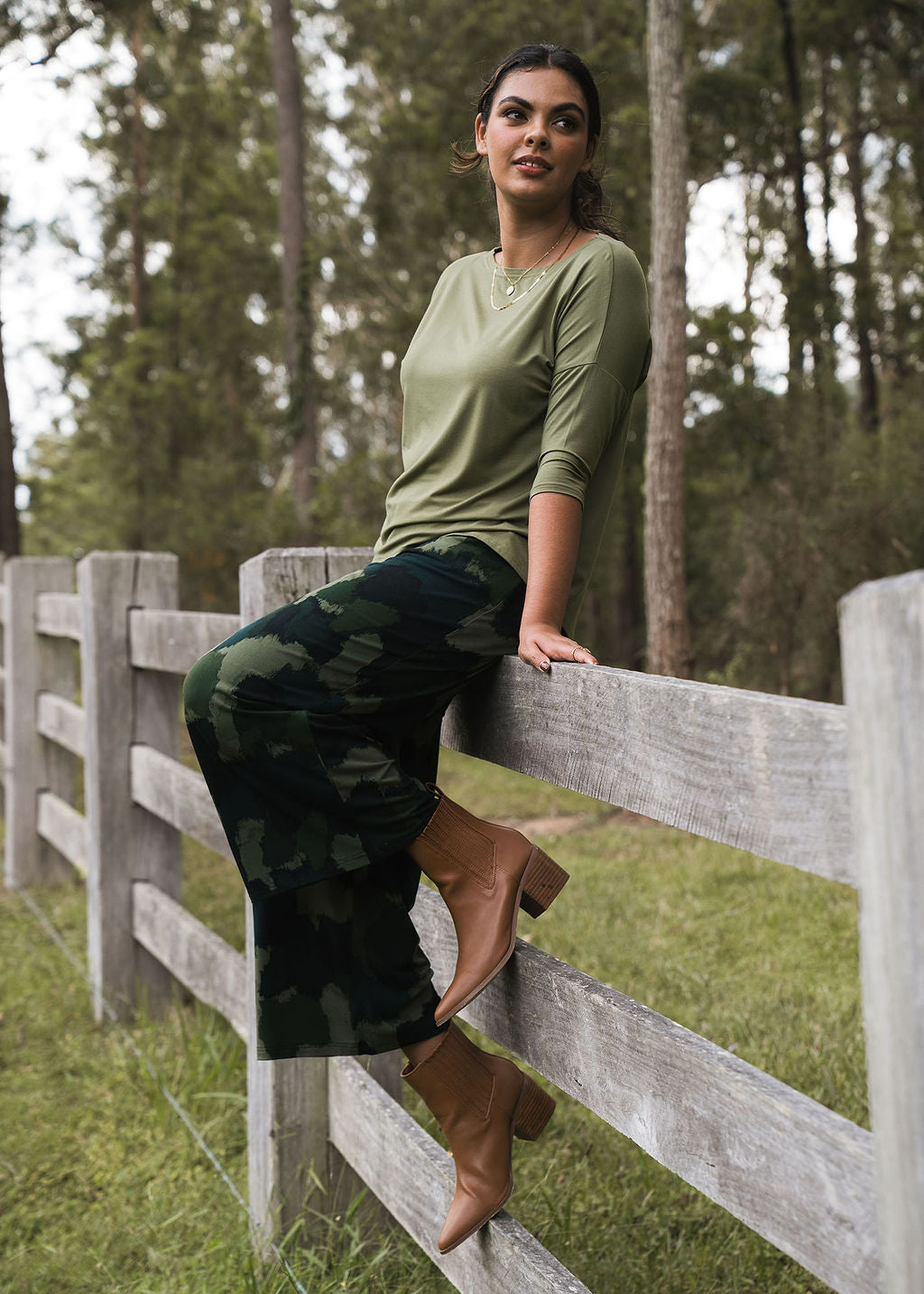 A woman in an olive green long-sleeve shirt and Ciara 7/8 Length Bamboo Culottes - Camouflage sits on a wooden fence in a grassy, wooded area with tan heeled boots, looking relaxed amid lush greenery and trees.
