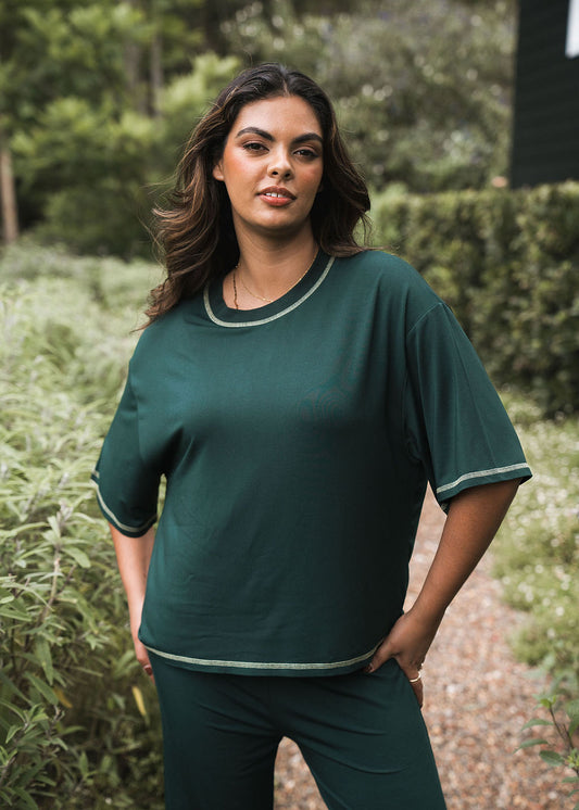 A woman with long dark hair models the Spears Bamboo T-Shirt in Dark Green, featuring light trim and a relaxed boxy fit, standing confidently on a gravel path surrounded by greenery with one hand on her hip.