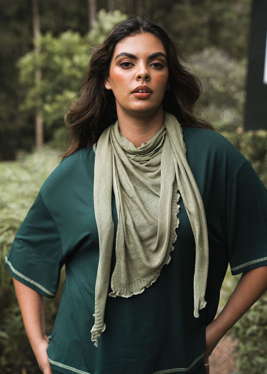 A woman with long dark hair stands outdoors, confidently wearing a loose dark green top and the Sassoon Cashmere/Bamboo Scarf in light green, with trees and foliage softly blurred in the background.