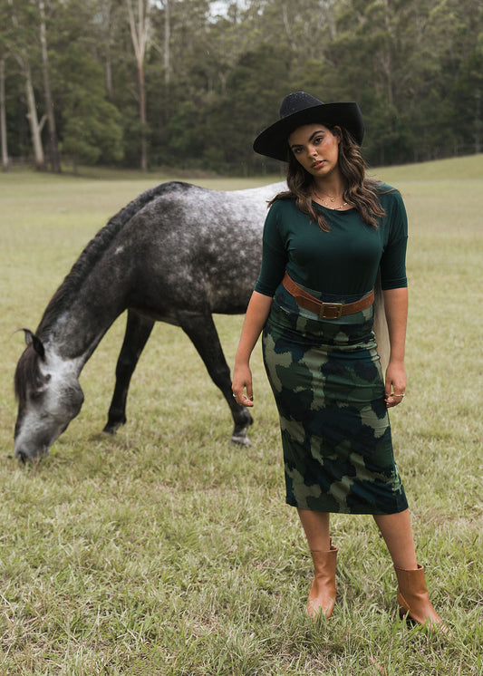 A woman wearing a dark green top, boots, black hat, and the Maxi Bamboo Whitney Tube Skirt with a brown belt stands on grass facing the camera. A gray dappled horse grazes behind her among trees.