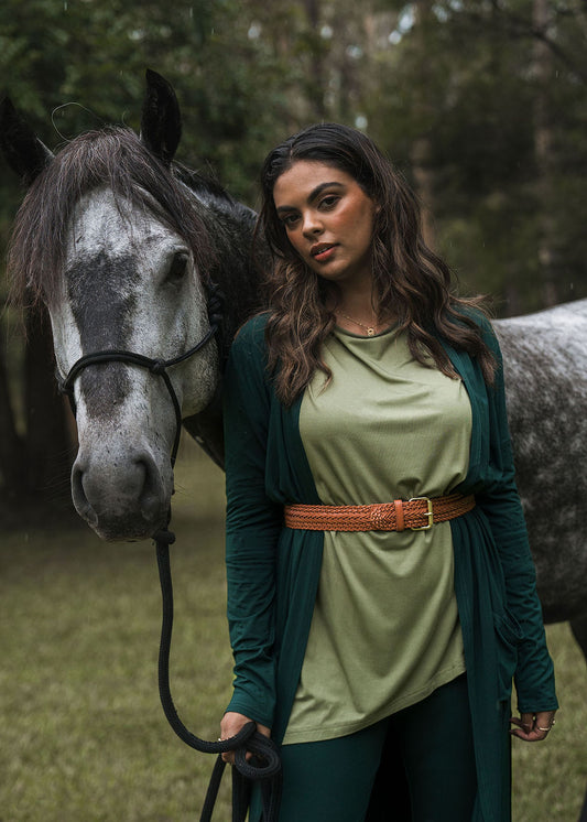 A woman in the Susie Oversized Asymmetrical Bamboo Top - Olive, styled with a brown belt, stands outdoors on grass beside a gray horse, holding its reins with trees in the background.