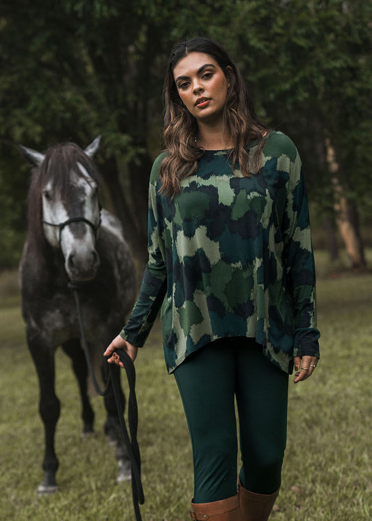 A woman stands outdoors on grass, wearing the Gina Long Sleeved Bamboo Top - Camouflage and dark green leggings. She holds a black lead attached to a dark gray horse, with trees in the background.