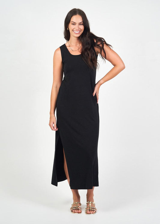 A woman smiles in the Janet Sleeveless Bamboo Maxi Dress - Black, featuring a side slit, paired with sandals and a necklace, against a plain white background.