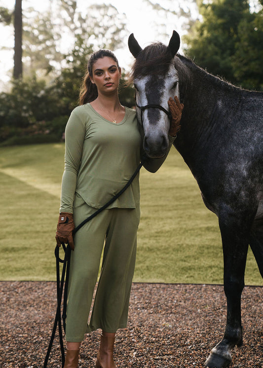 A woman wearing the Sia V Neck Bamboo Longsleeve Top in Olive Green stands on gravel, holding the reins of a dark gray horse and gently resting her gloved hand on its face, with trees and greenery in the background.