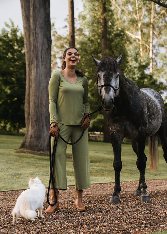 Wearing Ciara 7/8 Length Bamboo Culottes, a woman smiles outdoors as she holds the reins of a dark gray horse. A fluffy white cat sits at her feet, surrounded by green grass and sunlit trees.
