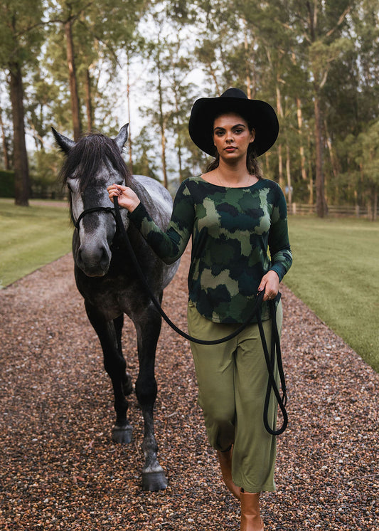 A woman in a black hat, olive pants, and the Adele Long Sleeve Bamboo Top in camouflage print walks barefoot on a gravel path, leading a black horse by its reins with trees and grass in the background.