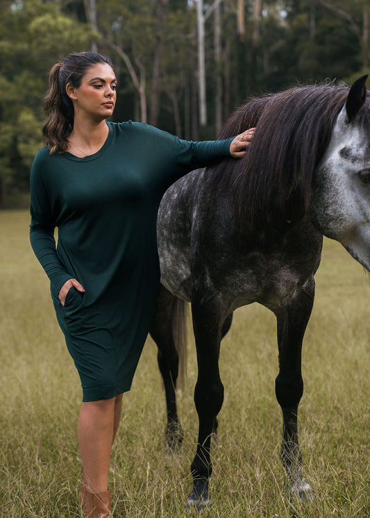 A woman wearing the Turner Long-Sleeve Bamboo T-Shirt Dress in dark green stands in a grassy field with one hand resting on a gray horse, trees in the background.