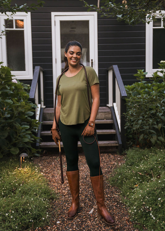 A woman in Luxe Bamboo Full-Length Legging, riding boots, an olive green shirt, and gloves stands on a gravel path before a dark house with green plants at the entrance, holding reins and a crop while smiling at the camera.