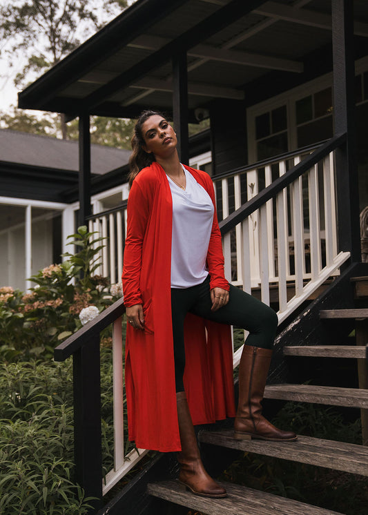 A woman on a porch steps confidently wears the Bamboo Longline Pocket Cardigan in Fiery Red, pairing it with dark leggings and brown knee-high boots. She stands before lush greenery and a dark house, embodying sustainable style.