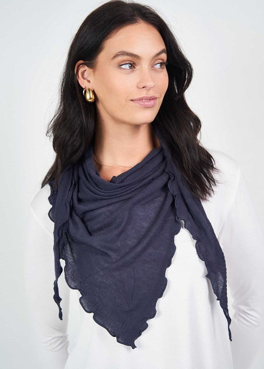 Woman with long dark hair wearing a white top, gold hoop earrings, and the Sassoon Cashmere/Bamboo Scarf in navy blue with scalloped edges, looking to her left against a light background.