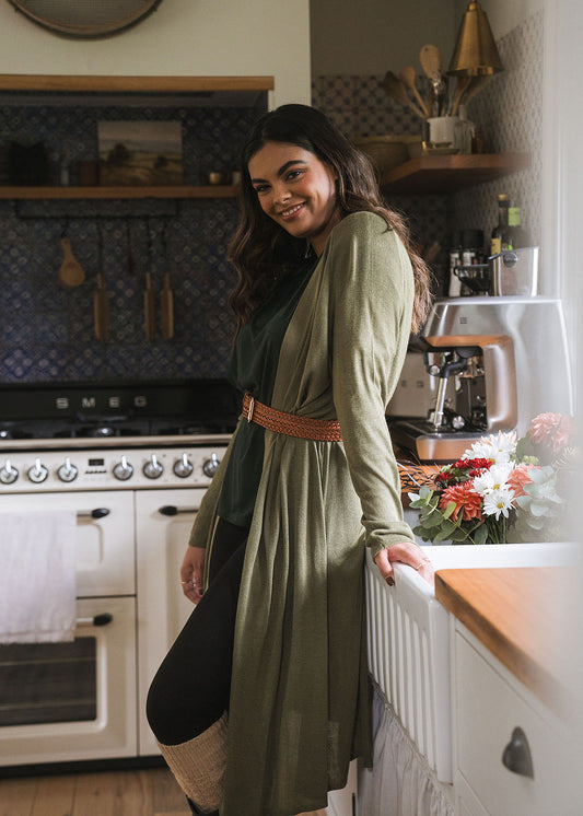 A woman with long dark hair, smiling at the camera, leans against a kitchen counter near flowers. She wears the Quatro Cashmere/Bamboo Duster Jacket in olive green and a brown belt. A stove and hanging utensils appear in the background.