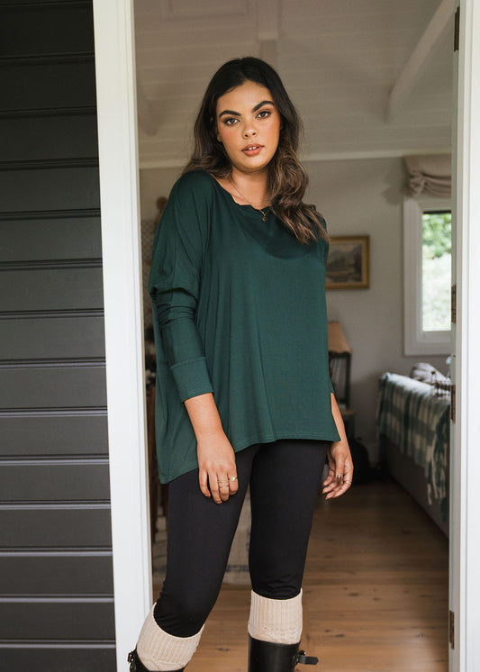 A woman with long hair stands in a cozy, well-lit room with wooden floors, wearing a Gina Long Sleeved Bamboo Top - Dark Green, black leggings, tan knit leg warmers over black boots.