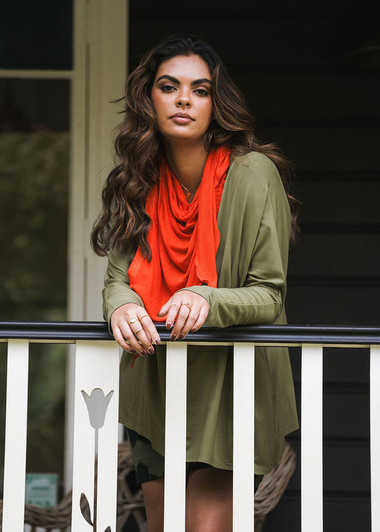 A woman with long wavy hair leans on a white porch railing, confidently wearing an olive green top, black shorts, and the Sassoon Cashmere/Bamboo Scarf in bright orange. A wicker chair and dark wall appear in the background.