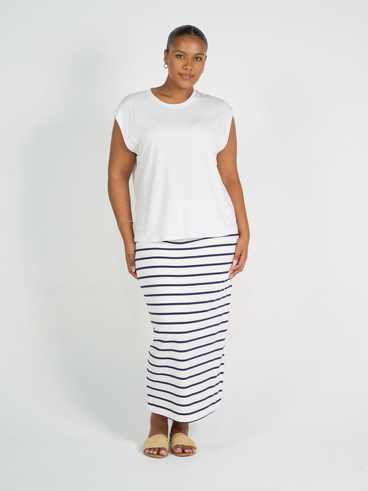 A woman stands against a white background wearing the Donna Cap Sleeve Bamboo Tee, a long white skirt with navy stripes, tan sandals, small hoop earrings, and a thin bracelet. She faces the camera with a neutral expression.