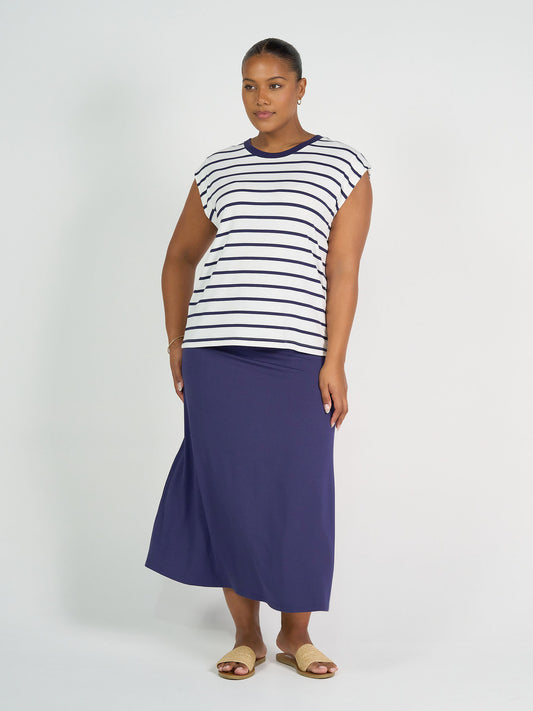 A woman stands against a plain background wearing the Donna Cap Sleeve Bamboo Tee in Blue & White Stripe, paired with a long navy skirt and beige slide sandals. Her hair is pulled back as she looks confidently at the camera—ideal for everyday style.