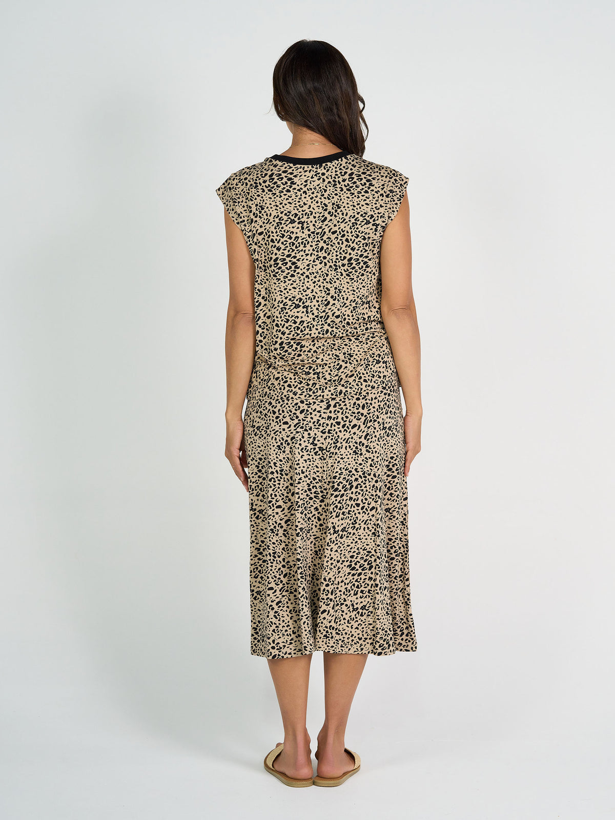 A woman faces away, wearing the Donna Cap Sleeve Bamboo Tee in brown leopard print, paired with flat sandals, against a plain white background.