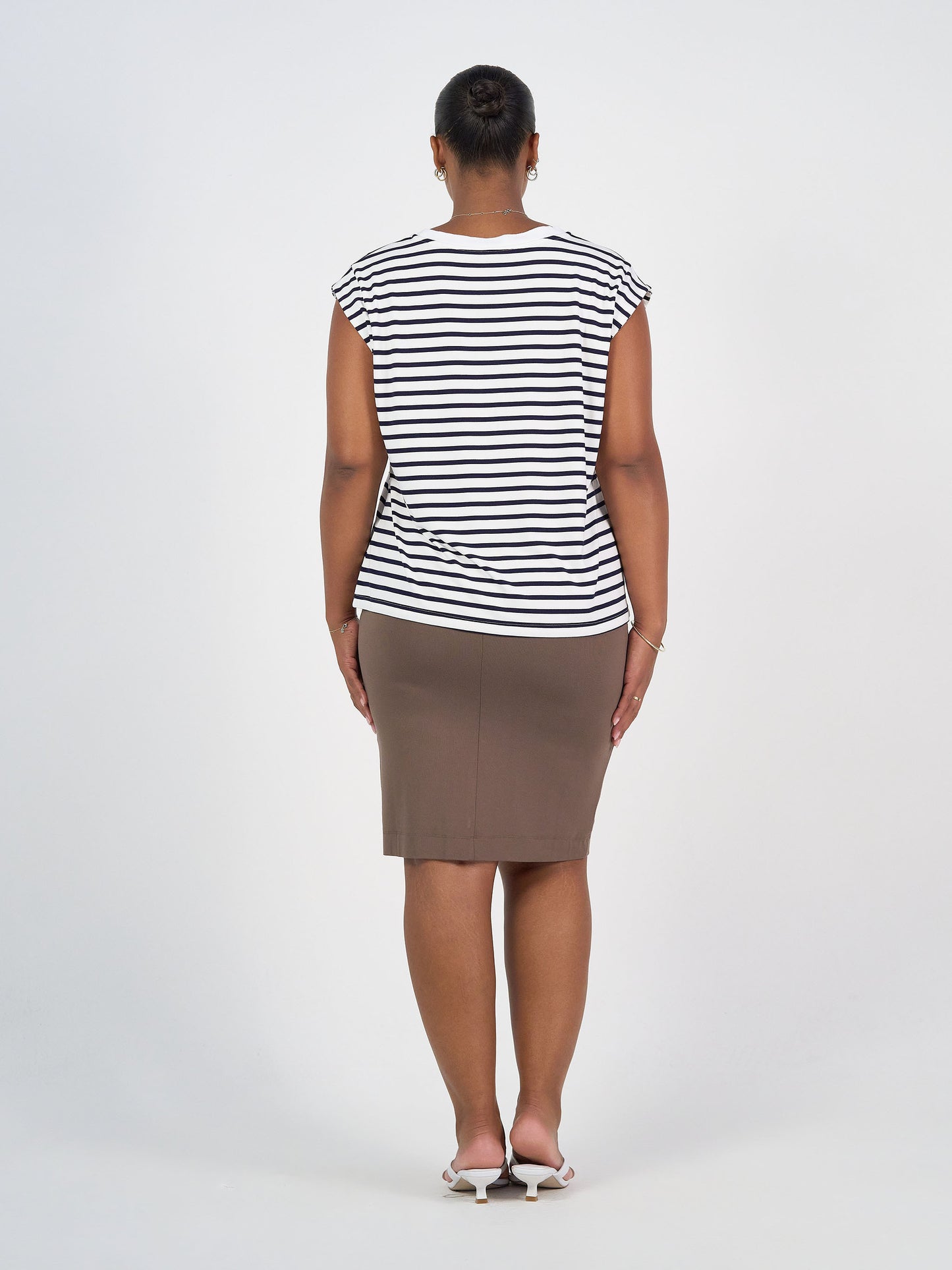 A woman stands facing away from the camera, wearing a navy and white striped Donna Cap Sleeve Bamboo Tee, a knee-length brown skirt, and white heeled sandals against a plain white background.